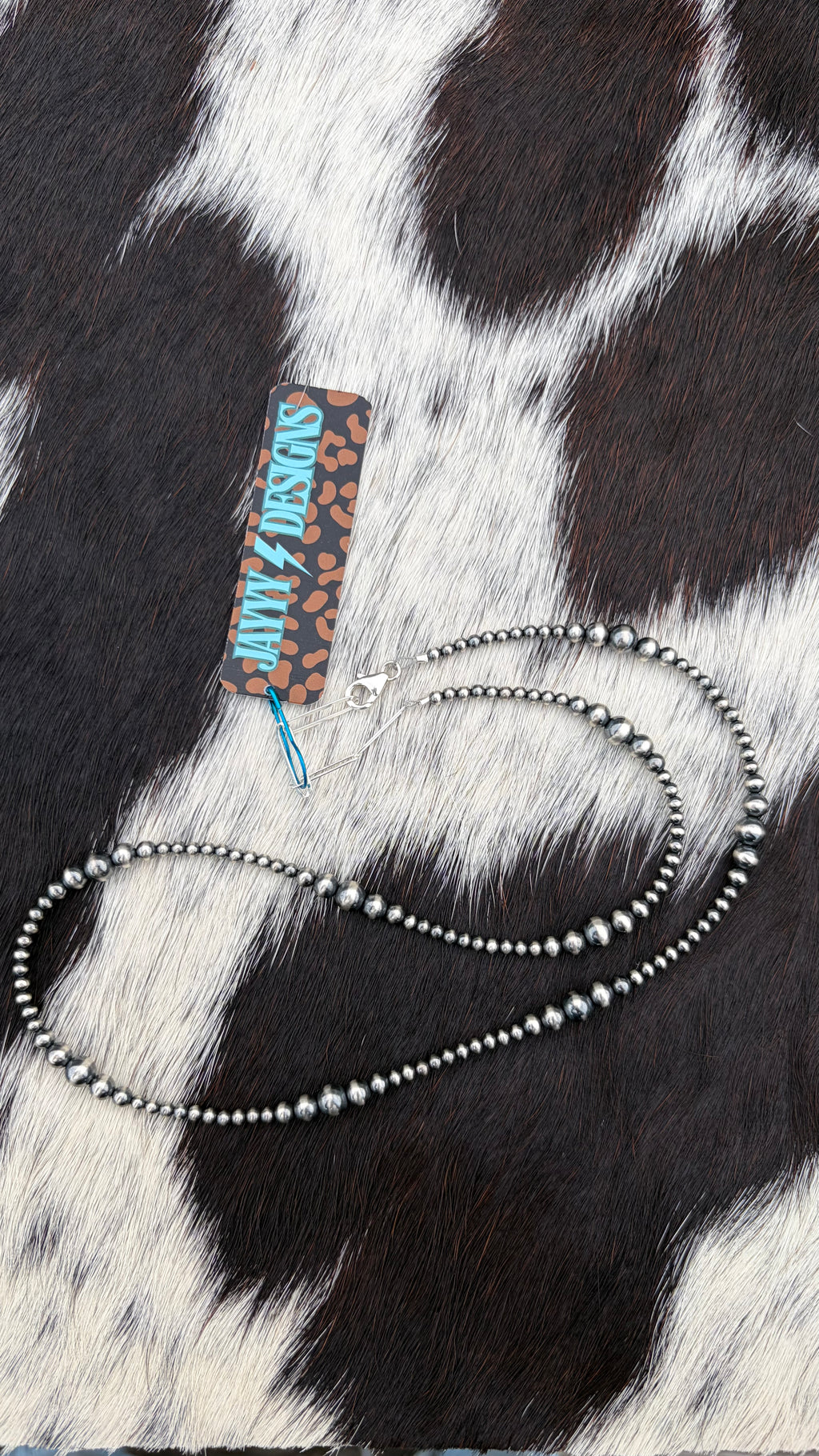 A sterling silver beaded necklace with small sterling silver beads, displayed on a black and white cowhide background next to a 'JayyyDesigns' brand tag.