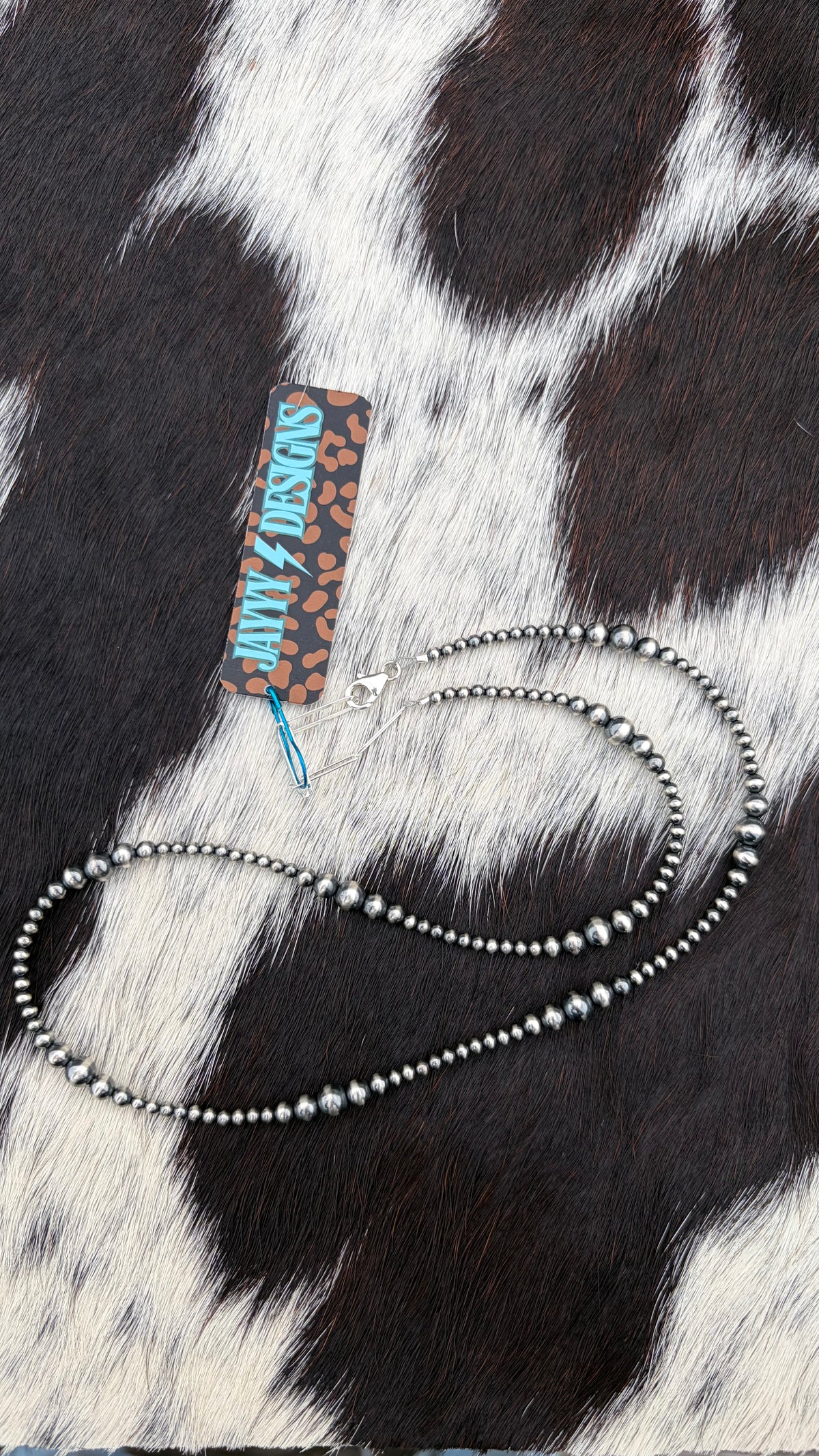 A sterling silver beaded necklace with small sterling silver beads, displayed on a black and white cowhide background next to a 'JayyyDesigns' brand tag.