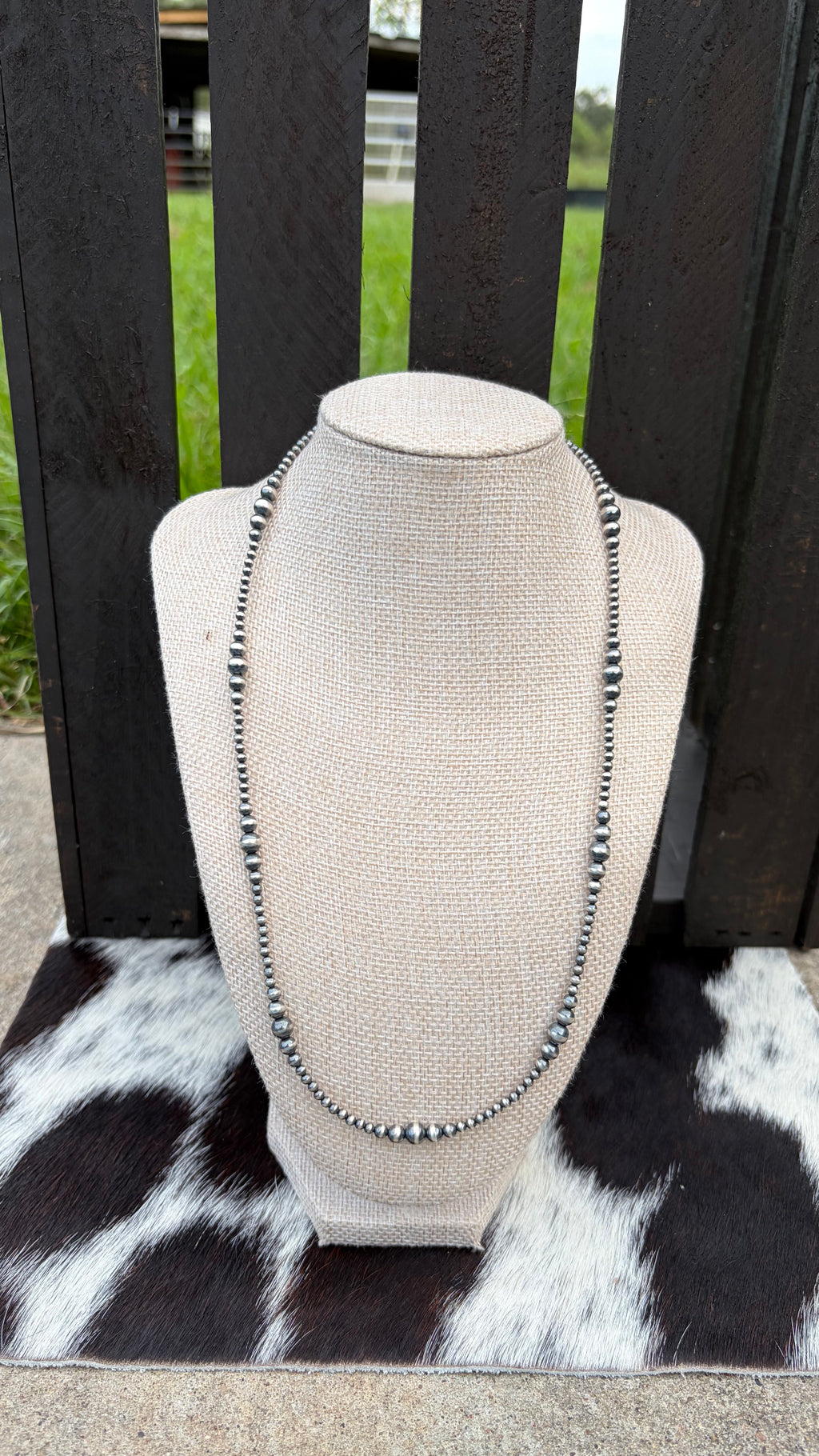 A sterling silver beaded necklace with round sterling silver beads and small sterling silver spacer beads, displayed on a beige fabric-covered jewelry bust. The bust is placed on a black-and-white cowhide rug in front of a dark wooden slat fence with grass visible in the background.