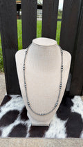 A sterling silver beaded necklace with round sterling silver beads and small sterling silver spacer beads, displayed on a beige fabric-covered jewelry bust. The bust is placed on a black-and-white cowhide rug in front of a dark wooden slat fence with grass visible in the background.