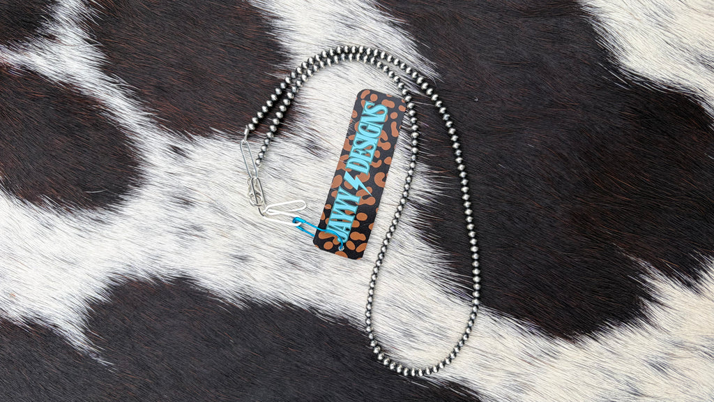 A 3mm sterling silver beaded necklace with a leopard print pattern in black and brown, with the text 'JAYYYDESIGNS' in blue and white. The necklace is displayed on a black and white cowhide background.