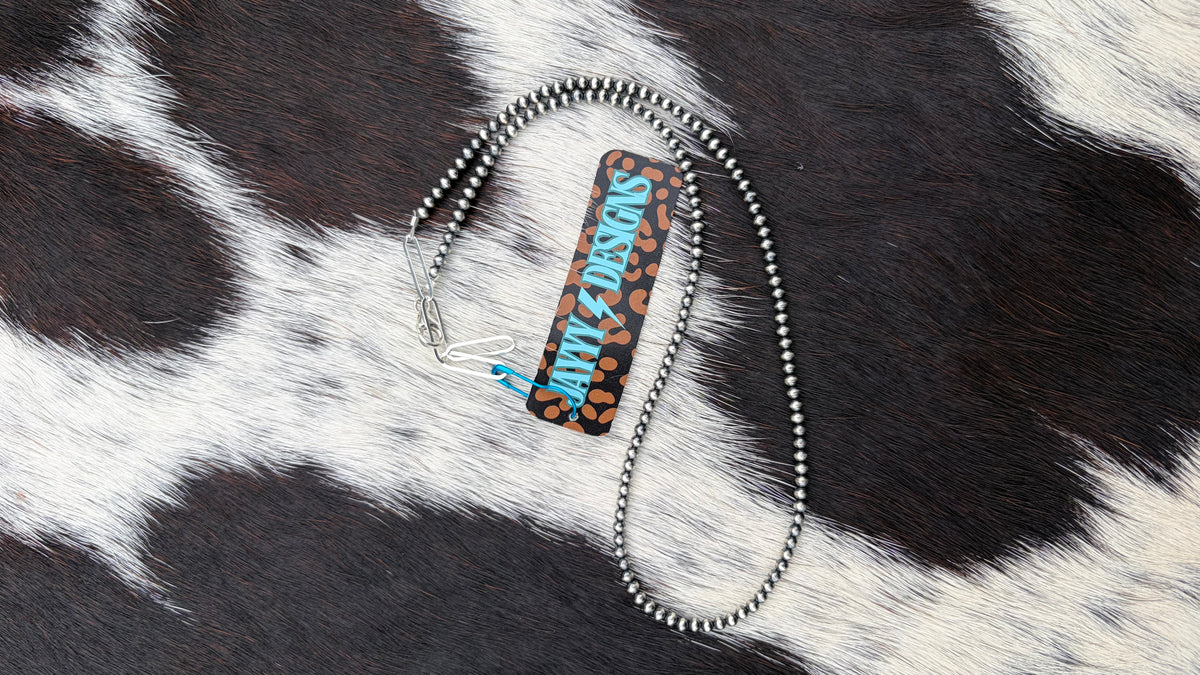 A 3mm sterling silver beaded necklace with a leopard print pattern in black and brown, with the text 'JAYYYDESIGNS' in blue and white. The necklace is displayed on a black and white cowhide background.