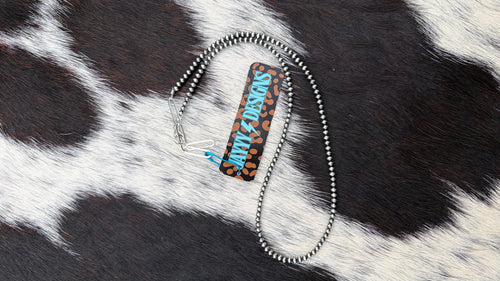 A 3mm sterling silver beaded necklace with a leopard print pattern in black and brown, with the text 'JAYYYDESIGNS' in blue and white. The necklace is displayed on a black and white cowhide background.