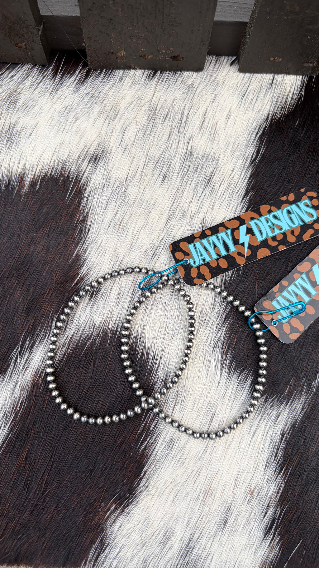 Two sterling silver 3mm stretch bracelets laid on a black-and-white cowhide surface, with branded paper tags attached that read JayyyDesigns.