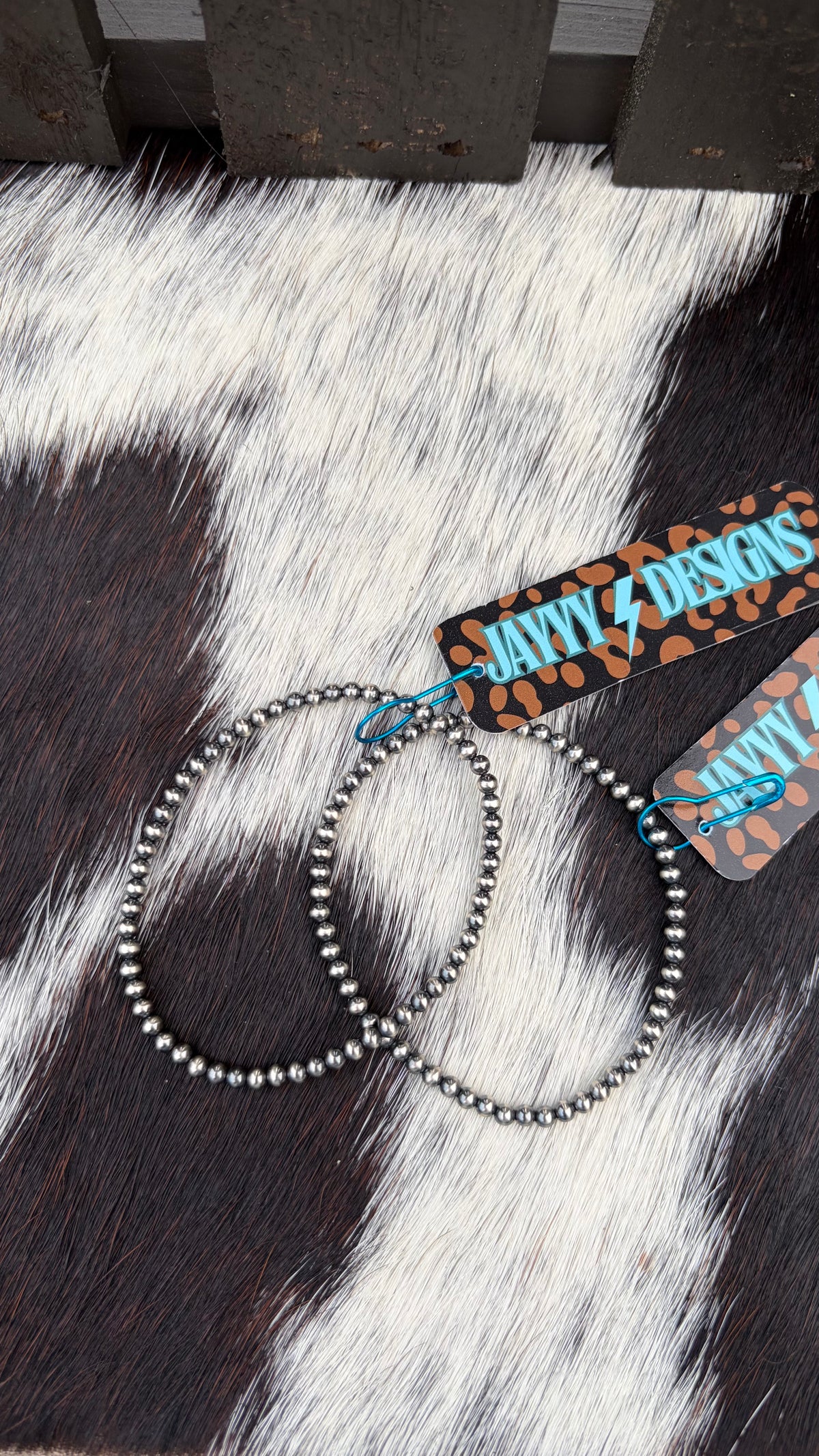 Two sterling silver 3mm stretch bracelets laid on a black-and-white cowhide surface, with branded paper tags attached that read JayyyDesigns.