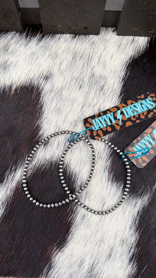 Two sterling silver 3mm stretch bracelets laid on a black-and-white cowhide surface, with branded paper tags attached that read JayyyDesigns.