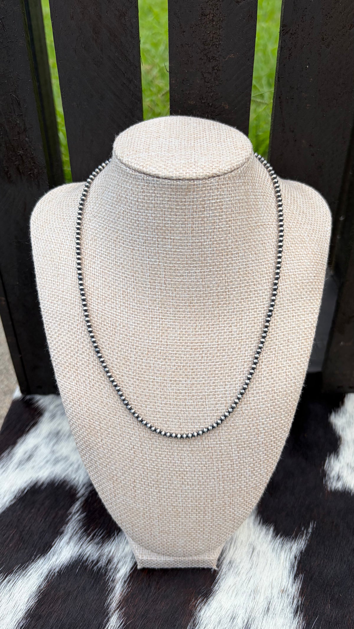 A 3mm sterling silver beaded necklace displayed on a beige woven jewelry bust, set against a dark wooden slatted background with a black-and-white cowhide surface below.