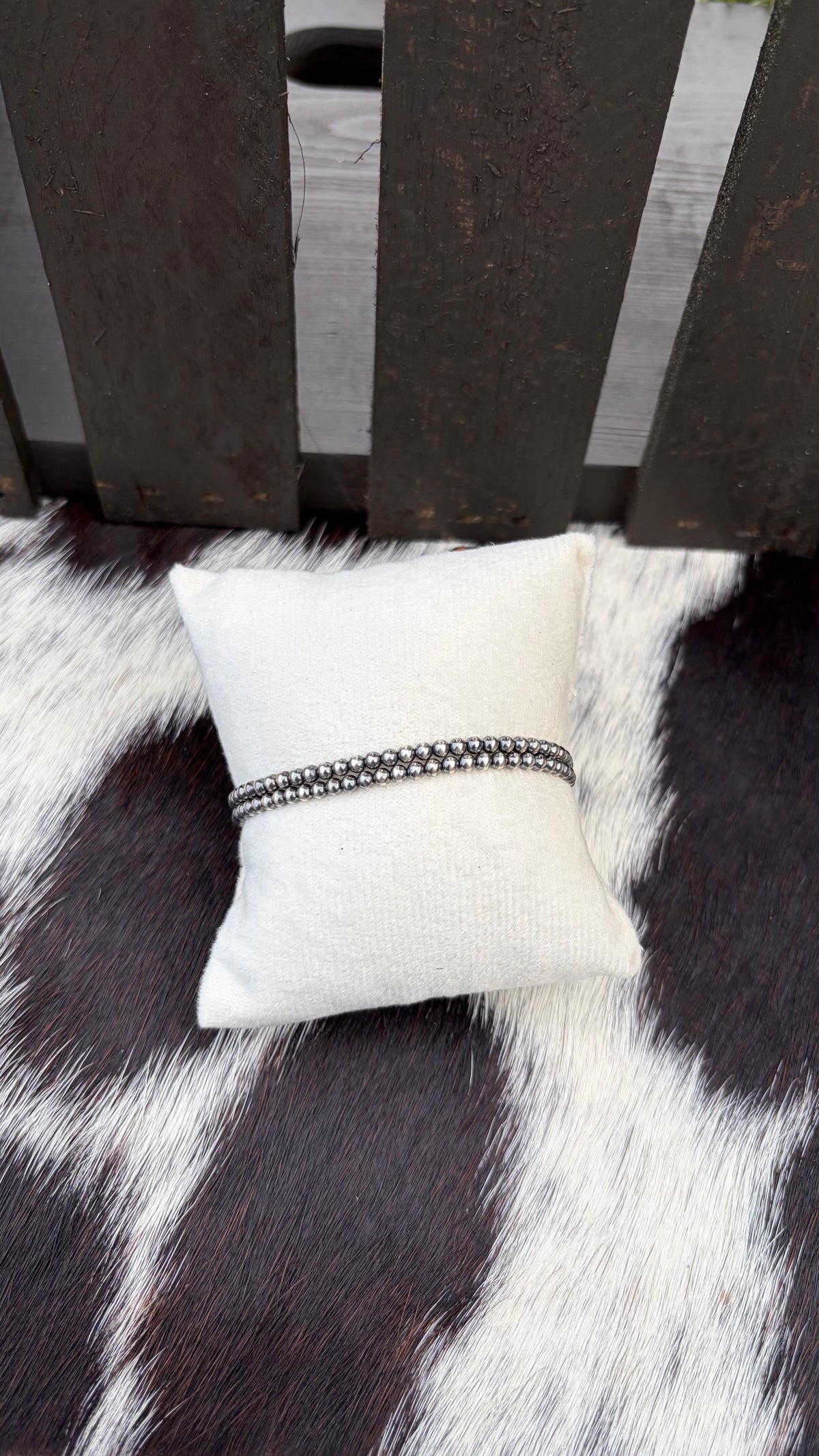 A cream bracelet pillow with  3 mm sterling silver stretch bracelet wrapped around the pillow, resting on a black and white cowhide rug in front of a dark wooden slatted backdrop.