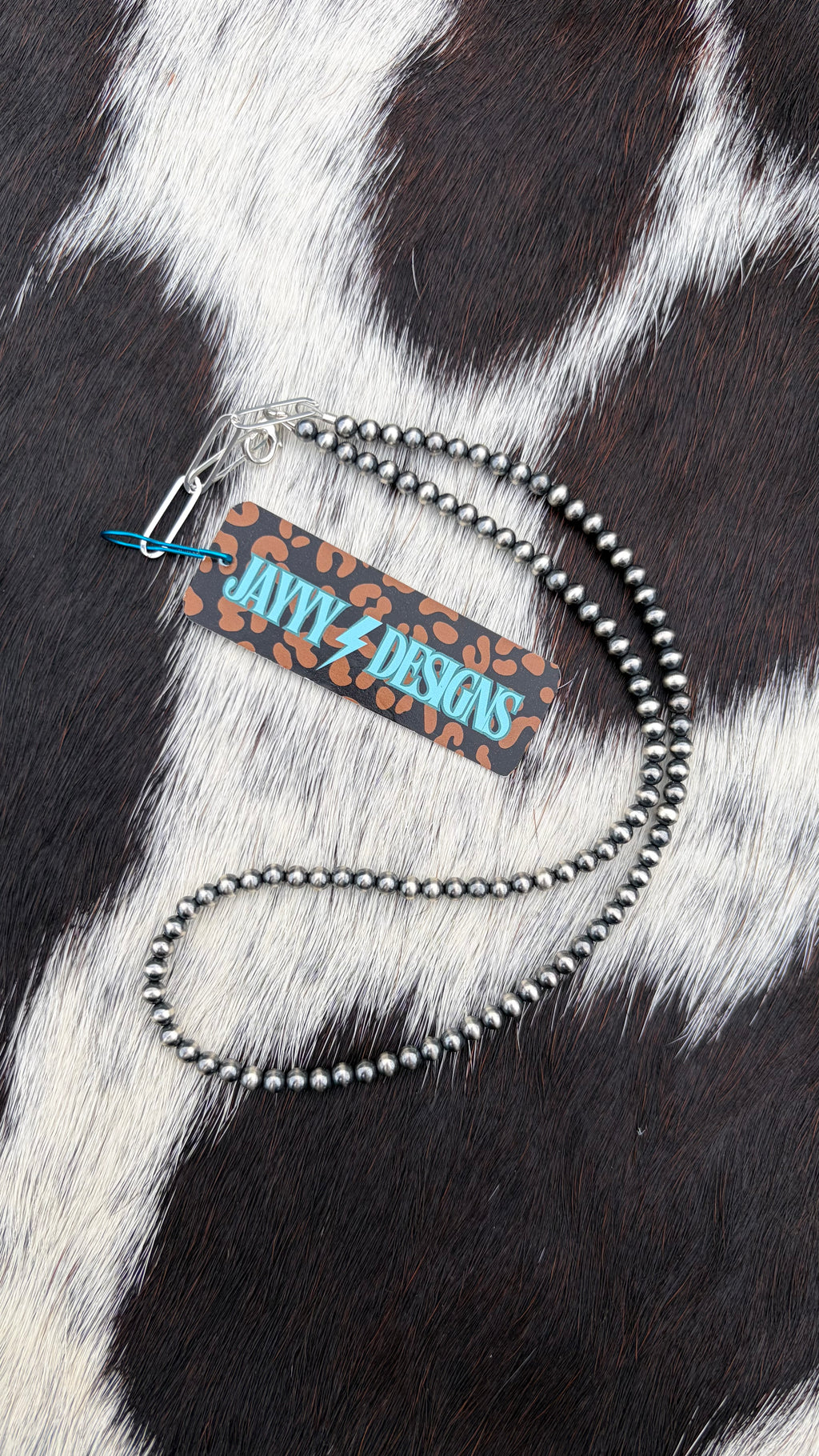 A 4mm sterling silver beaded necklace with a silver paperclip clasp and a branded paper tag, displayed on a black and white cowhide background.