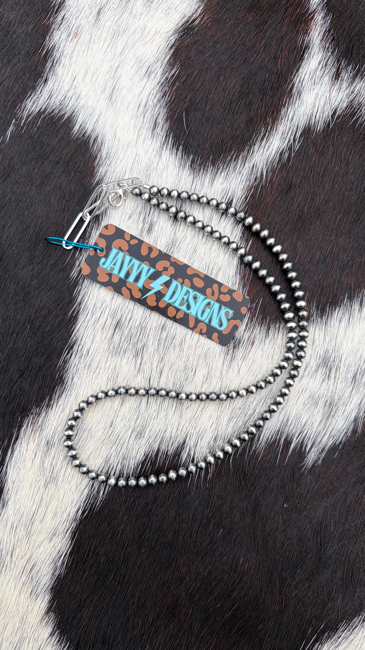 A 4mm sterling silver beaded necklace with a silver paperclip clasp and a branded paper tag, displayed on a black and white cowhide background.