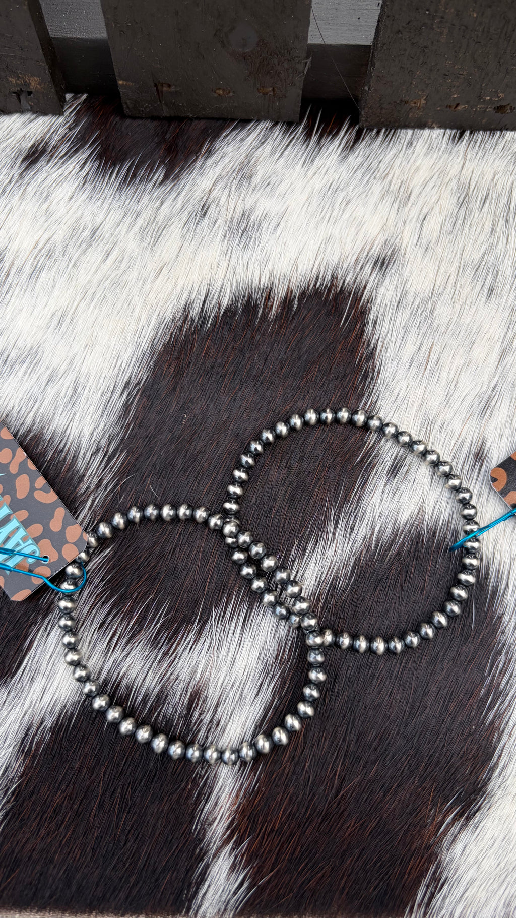 Two 4mm beaded hoop bracelets with dark metallic beads, displayed on a black and white cowhide surface with a wooden fence in the background.
