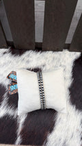 A 4mm beaded silver bracelet displayed on a white fabric pillow, set on a black and white cowhide rug in front of a dark wooden slatted surface.