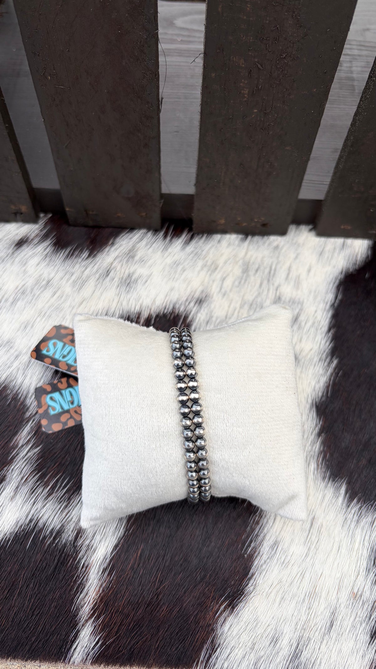 A 4mm beaded silver bracelet displayed on a white fabric pillow, set on a black and white cowhide rug in front of a dark wooden slatted surface.