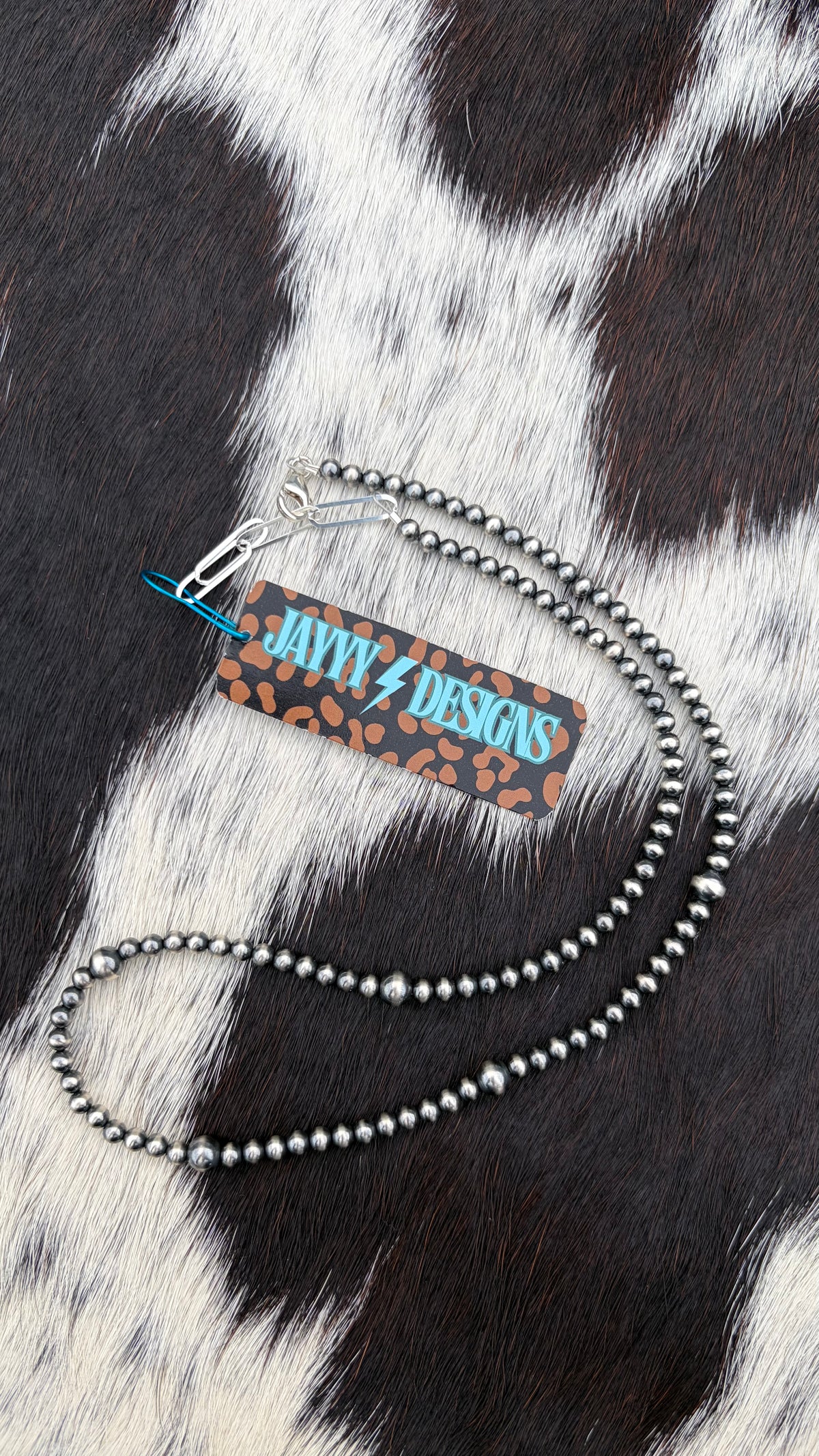 A 4mm and 6mm sterling silver beaded necklace with a lobster clasp and a rectangular brand tag reading 'JAYYYDESIGNS' in turquoise on a leopard-print background, laid flat on a black-and-white cowhide surface.