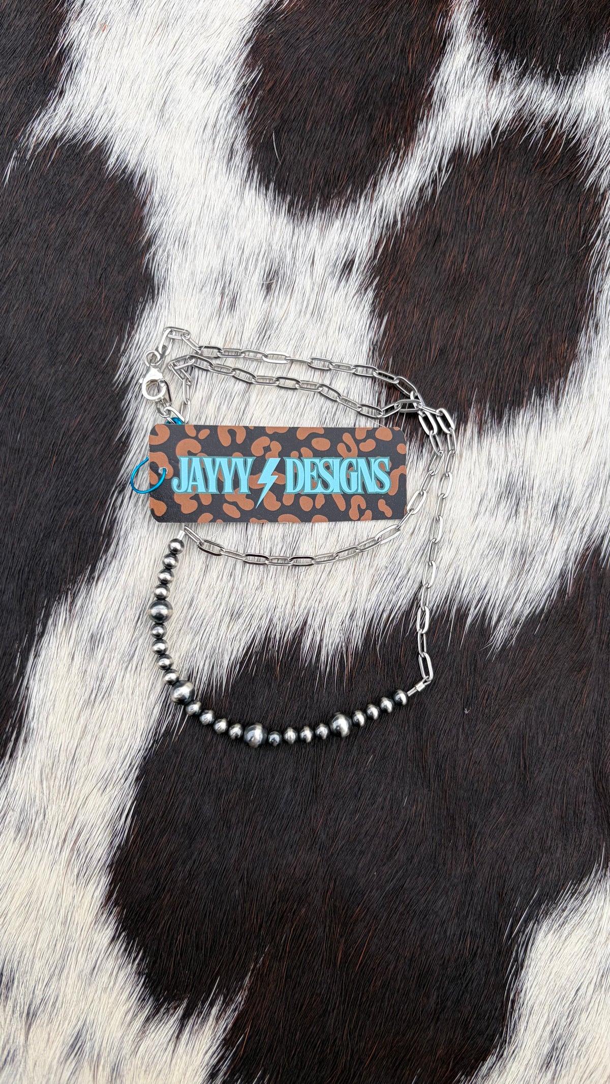 A stainless steel chain 4mm and 6mm beaded bar necklace with a rectangular leopard-print charm featuring the text 'JAYYYDESIGNS' in blue and a brown leopard-print price tag, displayed on a black and white cowhide background.
