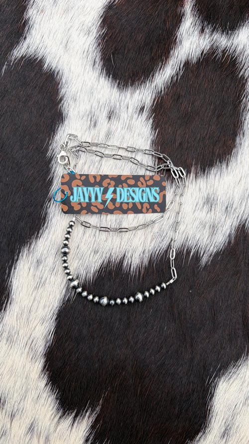 A stainless steel chain 4mm and 6mm beaded bar necklace with a rectangular leopard-print charm featuring the text 'JAYYYDESIGNS' in blue and a brown leopard-print price tag, displayed on a black and white cowhide background.