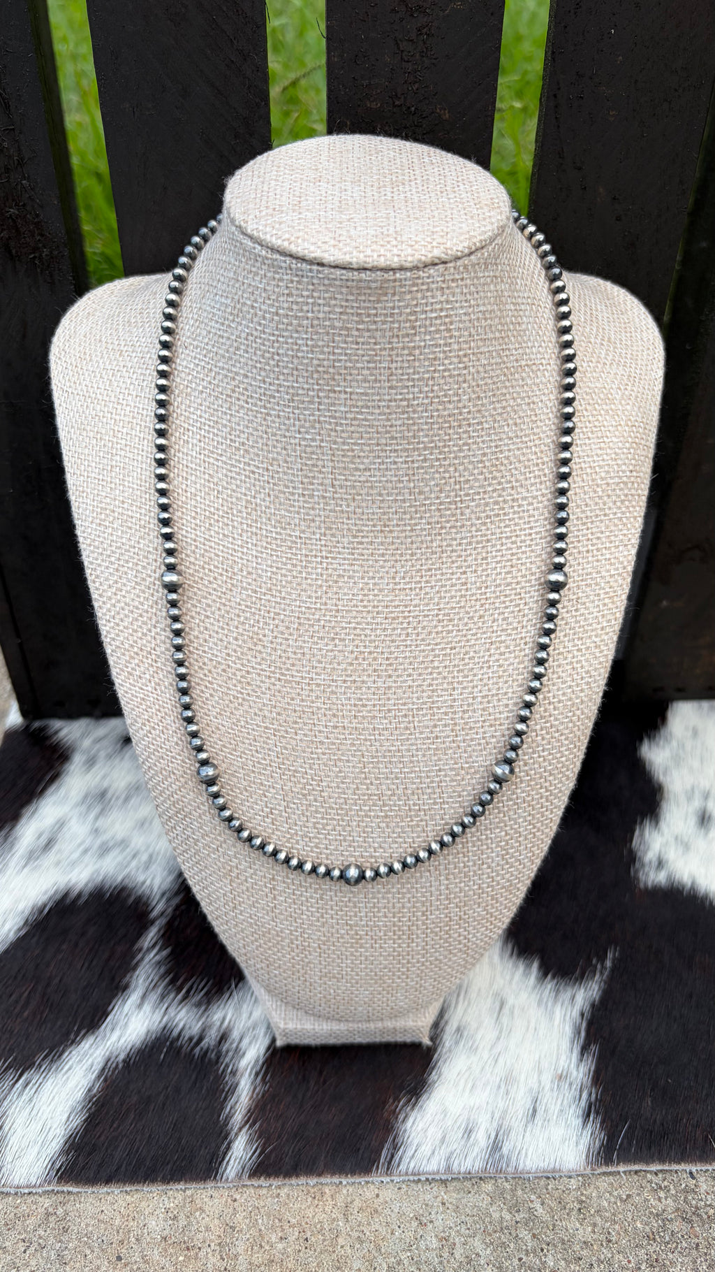 A 4mm and 6mm sterling silver beaded necklace with sterling silver round beads displayed on a textured beige fabric jewelry bust. The bust is set against a dark wooden slatted background with a black and white cowhide rug underneath.