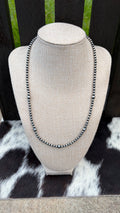 A 4mm and 6mm sterling silver beaded necklace with sterling silver round beads displayed on a textured beige fabric jewelry bust. The bust is set against a dark wooden slatted background with a black and white cowhide rug underneath.