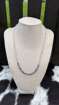 A 4mm and 6mm sterling silver beaded bar necklace with a stainless steel chain displayed on a beige fabric jewelry bust, set against a dark wooden slat background with a black-and-white cowhide surface below.