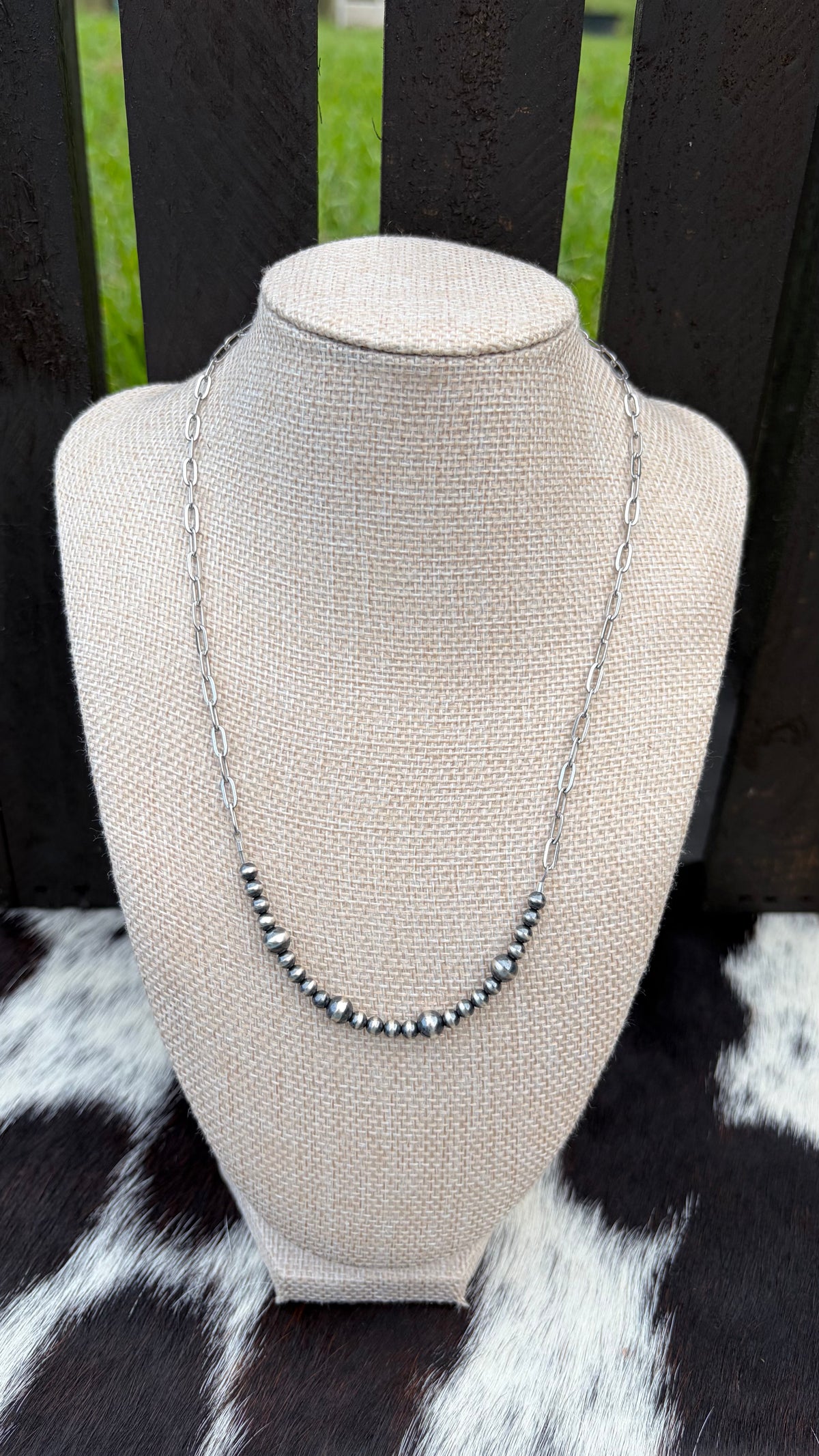 A 4mm and 6mm sterling silver beaded bar necklace with a stainless steel chain displayed on a beige fabric jewelry bust, set against a dark wooden slat background with a black-and-white cowhide surface below.