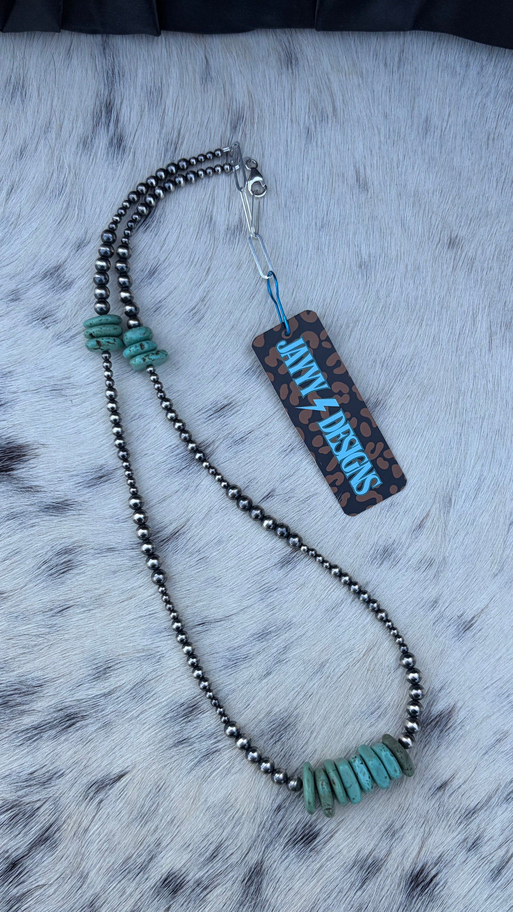 A beaded necklace with dark metallic beads, turquoise-colored stone accents, and a matching rectangular tag reading 'JAYYYDESIGNS' is displayed on a white faux fur surface.
