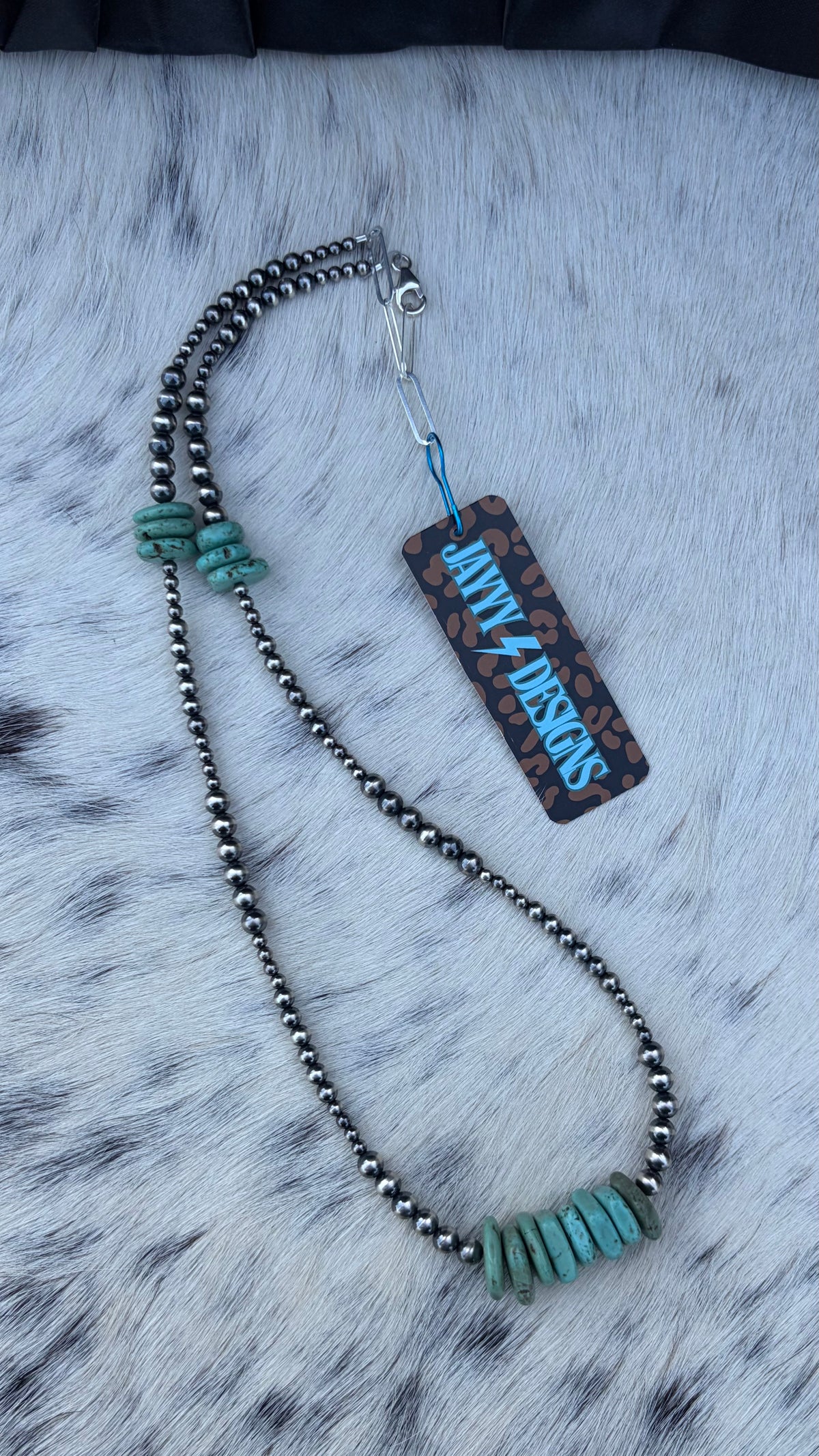 A beaded necklace with dark metallic beads, turquoise-colored stone accents, and a matching rectangular tag reading 'JAYYYDESIGNS' is displayed on a white faux fur surface.