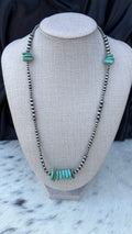 A beaded necklace displayed on a textured fabric jewelry bust. The necklace features dark metallic beads with a central cluster of turquoise-colored stone beads and matching turquoise accents at the top.