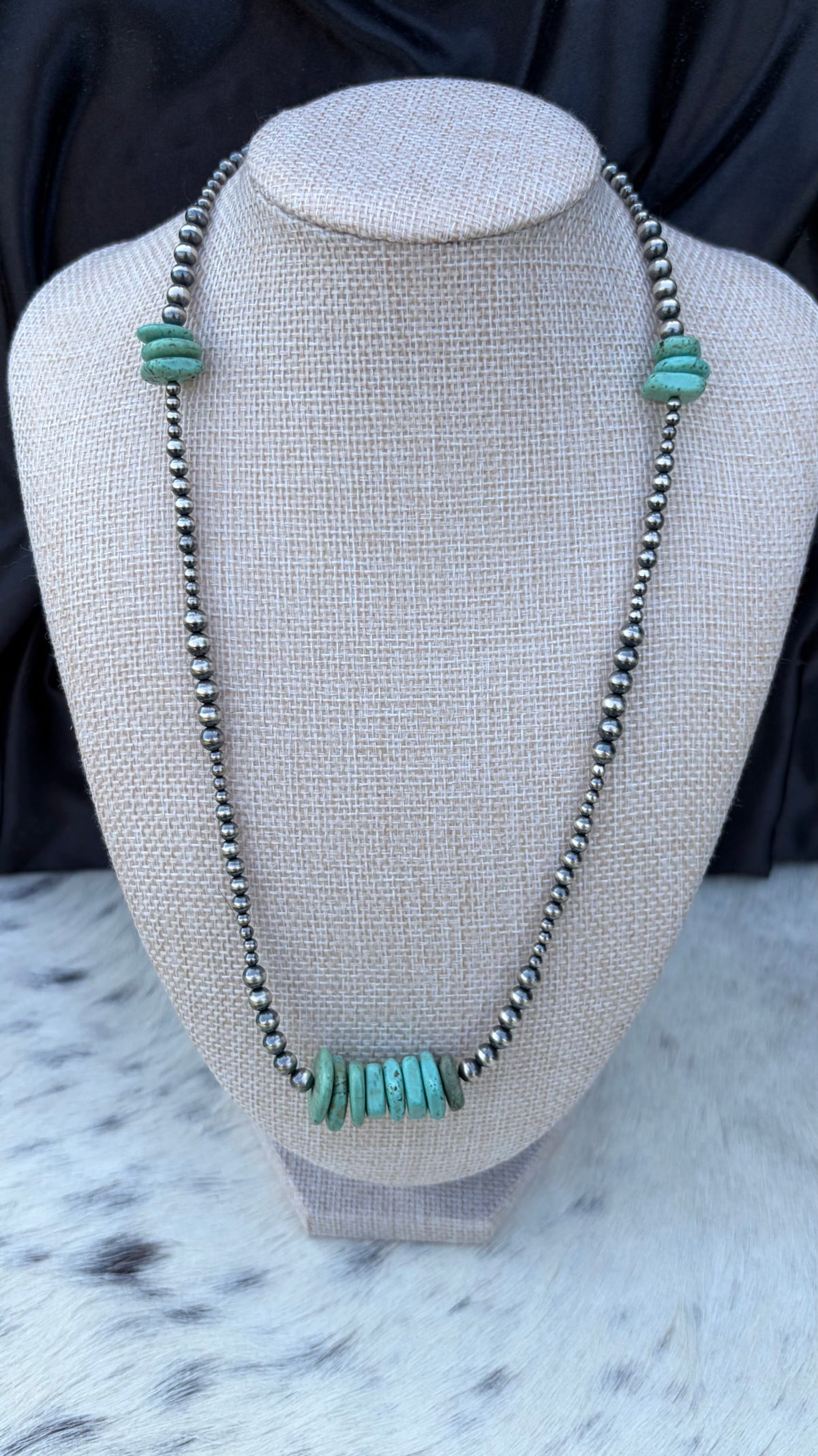 A beaded necklace displayed on a textured fabric jewelry bust. The necklace features dark metallic beads with a central cluster of turquoise-colored stone beads and matching turquoise accents at the top.